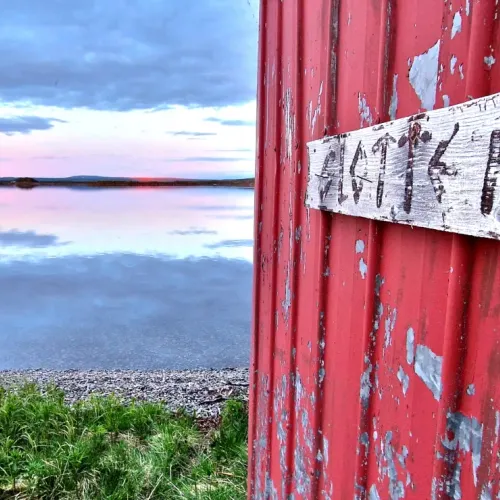 Rödmålad bod med bleknad skylt vid sjökanten