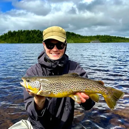 Man med solglasögon håller öring framför fjällsjö