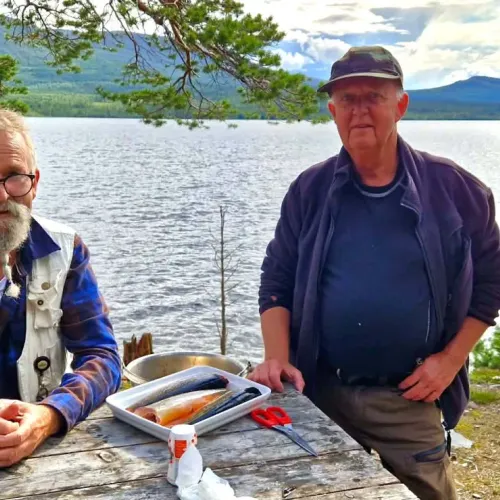 Två män rensar fisk vid bord utmed sjön