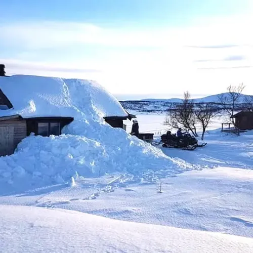 Snödriva mot stugväggen, skoter i bakgrunden